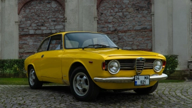Klassischer gelber Alfa Romeo, geparkt auf Kopfsteinpflaster vor historischen Steinmauern.