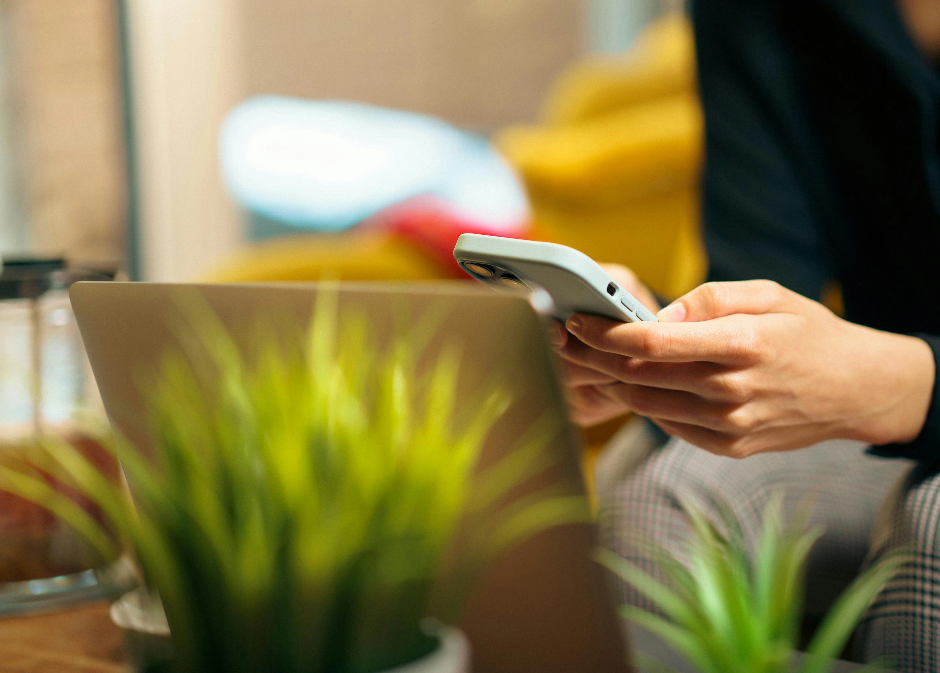 Eine Frau erledigt Multitasking auf ihrem Smartphone und Laptop, umgeben von grünen Pflanzen in einem gemütlichen Wohnzimmer.