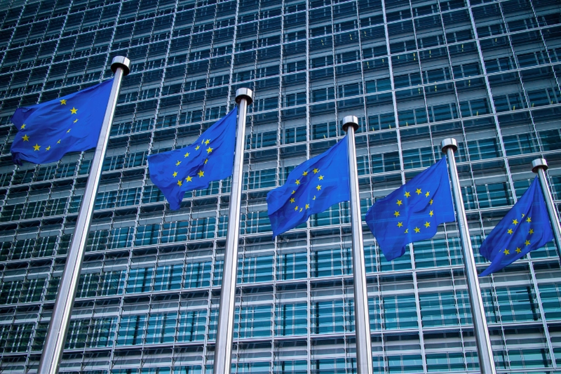 Fünf Flaggen der Europäischen Union wehen vor einem modernen Gebäude mit Glasfassade. Die Flaggen zeigen den blauen Hintergrund mit gelben Sternen, charakteristisch für die EU.