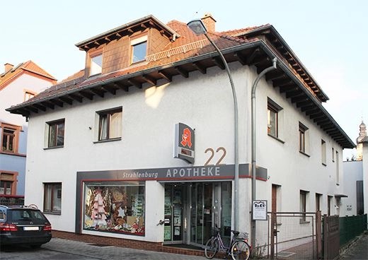 Vorderansicht einer Apotheke in einem zweistöckigen Wohn- und Geschäftsgebäude, Schaufenster mit Dekoration, Hausnummer 22 und abgestelltes Fahrrad davor.