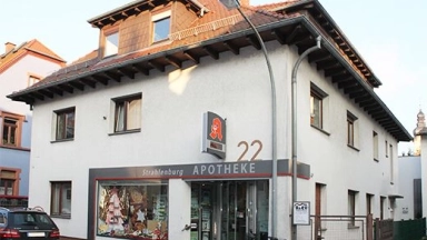 Vorderansicht einer Apotheke in einem zweistöckigen Wohn- und Geschäftsgebäude, Schaufenster mit Dekoration, Hausnummer 22 und abgestelltes Fahrrad davor.