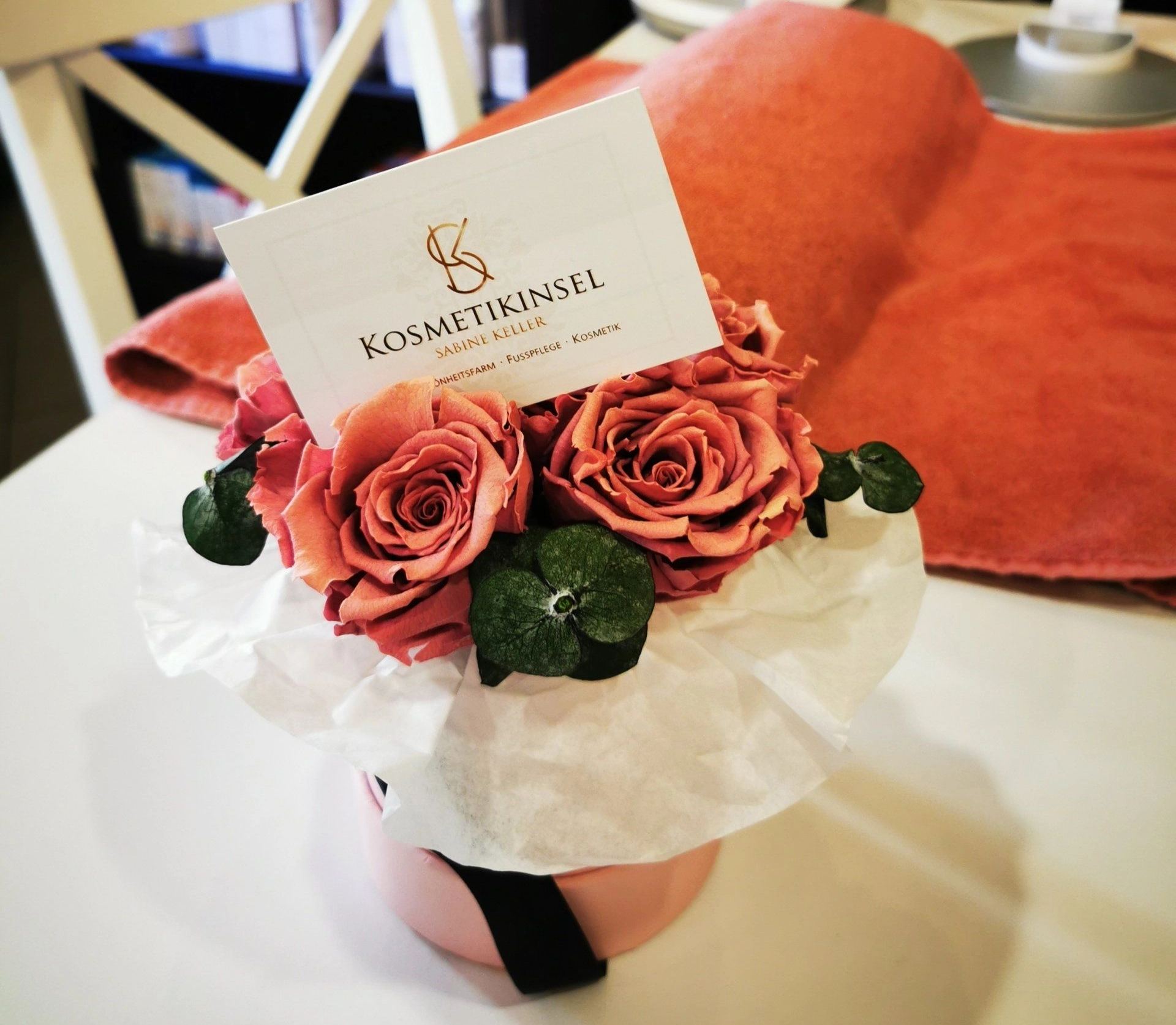 Rosa Rosen in einer runden Geschenkbox mit weißem Papier, ergänzt durch grüne Blätter und eine Visitenkarte eines Kosmetikstudios.