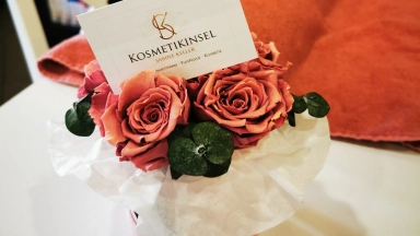 Rosa Rosen in einer runden Geschenkbox mit weißem Papier, ergänzt durch grüne Blätter und eine Visitenkarte eines Kosmetikstudios.