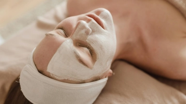 Frau mit geschlossenen Augen auf Liege, trägt Stirnband und weiße Gesichtsmaske.