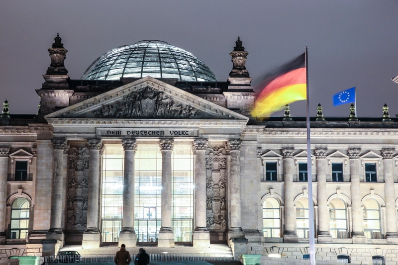 Das Bild zeigt die Reichstagsgebäude in Berlin bei Nacht. Im Vordergrund wehen die deutsche und die europäische Flagge. Die Fassade des Gebäudes ist mit beeindruckenden architektonischen Details verziert, darunter große Fenster und eine Kuppel.
