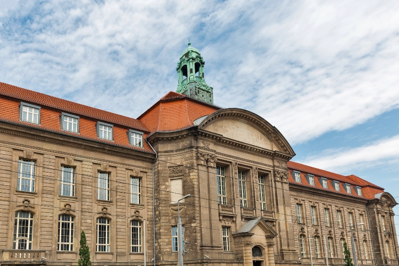 Historisches Gebäude mit herausragender Architektur, einschließlich einem großen Eingang und einem Glockenturm. Die Fassade ist mit symmetrischen Fenstern und einem roten Ziegeldach gestaltet. Der Himmel zeigt einige Wolken.