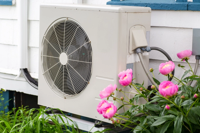 Klimaanlage an einer Außenwand, umgeben von rosa Pfingstrosen. Die Einheit hat einen großen Ventilator und ist in einem freundlichen, gepflegten Garten platziert.