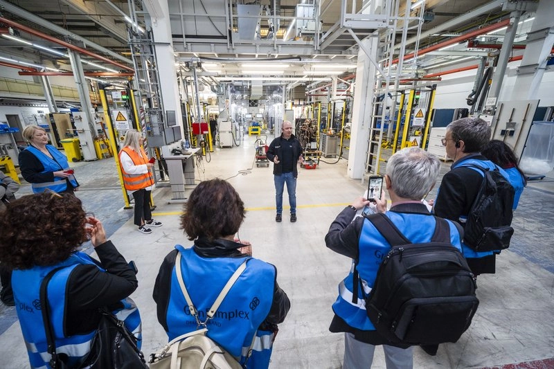 Eine Gruppe von Personen in blauen Westen steht in einer Fabrikhalle. Ein Mann in schwarzer Kleidung spricht zu ihnen, während einige der Anwesenden Notizen machen oder Fotos mit ihren Handys aufnehmen. Im Hintergrund sind industrielle Maschinen und Ausrüstungen zu sehen.