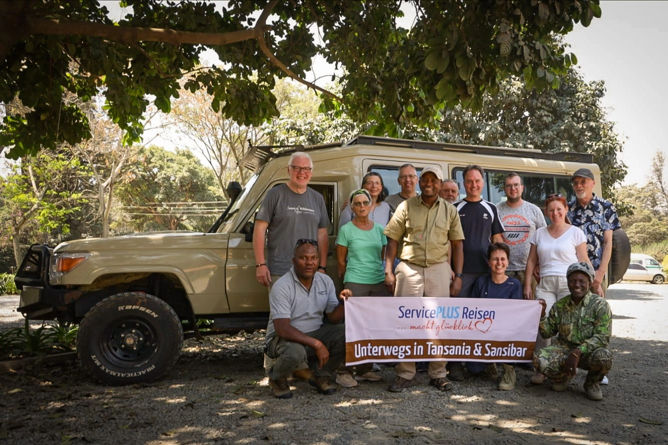 Gruppe von Erwachsenen vor einem Safari-Geländewagen, einige halten ein Banner mit der Aufschrift ‚Unterwegs in Tansania & Sansibar‘.