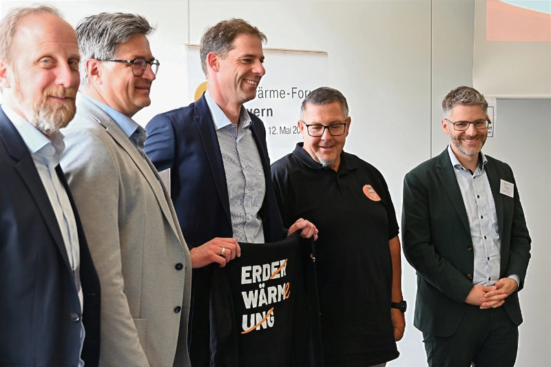 Fünf Männer stehen nebeneinander und lächeln. Sie halten ein schwarzes T-Shirt mit der Aufschrift "ERDERWÄRMUNG". Im Hintergrund befindet sich ein Banner mit dem Titel einer Veranstaltung. Die Umgebung wirkt professionell und förmlich.