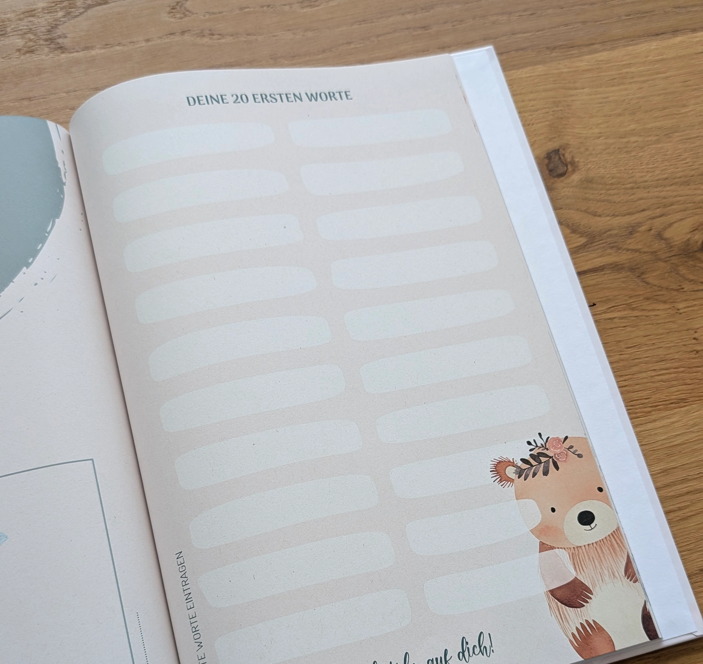 Aufgeschlagene Babybuch-Seite mit dem Titel „Deine 20 ersten Worte“, leeren Eintragsfeldern und einer kleinen Bären-Illustration auf einem Holztisch.