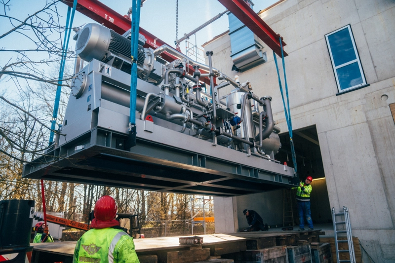 Ein großer Maschinenblock wird mit einem Kran an ein Gebäude angehoben. Arbeiter in Schutzkleidung und Helmen sind damit beschäftigt, die Maschine zu installieren oder zu sichern. Im Hintergrund sind Bäume und Baugerüste sichtbar.