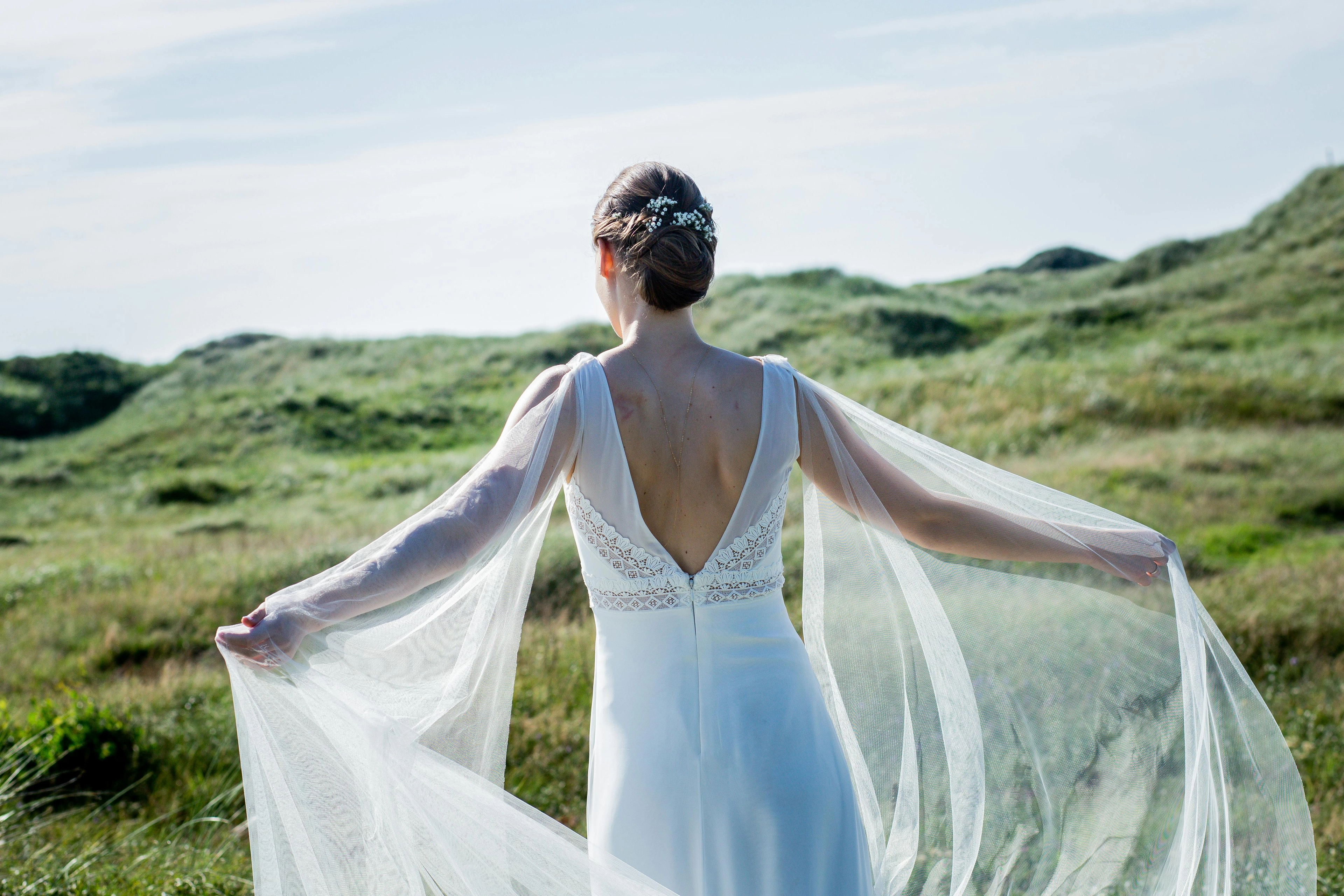 Rückansicht einer Frau in weißem Brautkleid mit fließenden, transparenten Ärmeln vor grünen Dünen.