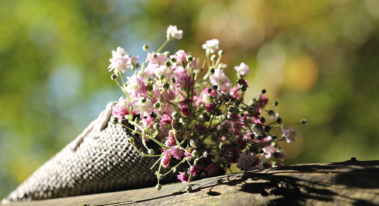 Kleiner Strauß aus rosa-weißen Blüten in grobem Tuch auf einem Holzstamm vor unscharf grünem Hintergrund.