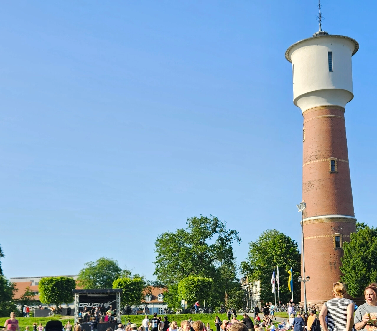 Menschenmenge bei einer Freiluftveranstaltung auf einer Wiese neben einem hohen Backstein-Wasserturm unter klarem Himmel.