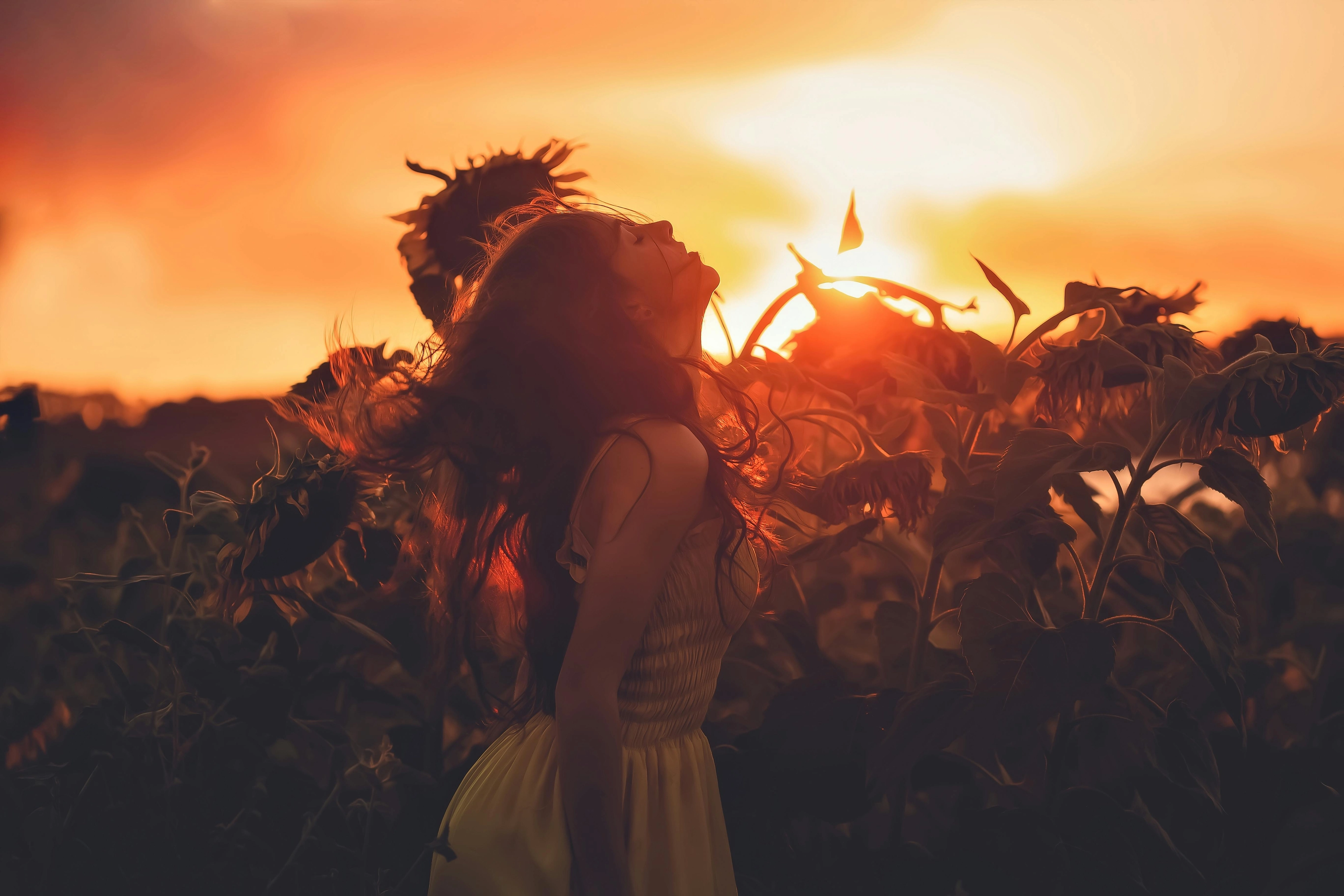 Mädchen in gelbem Kleid steht zwischen Sonnenblumen und blickt bei Sonnenuntergang nach oben.