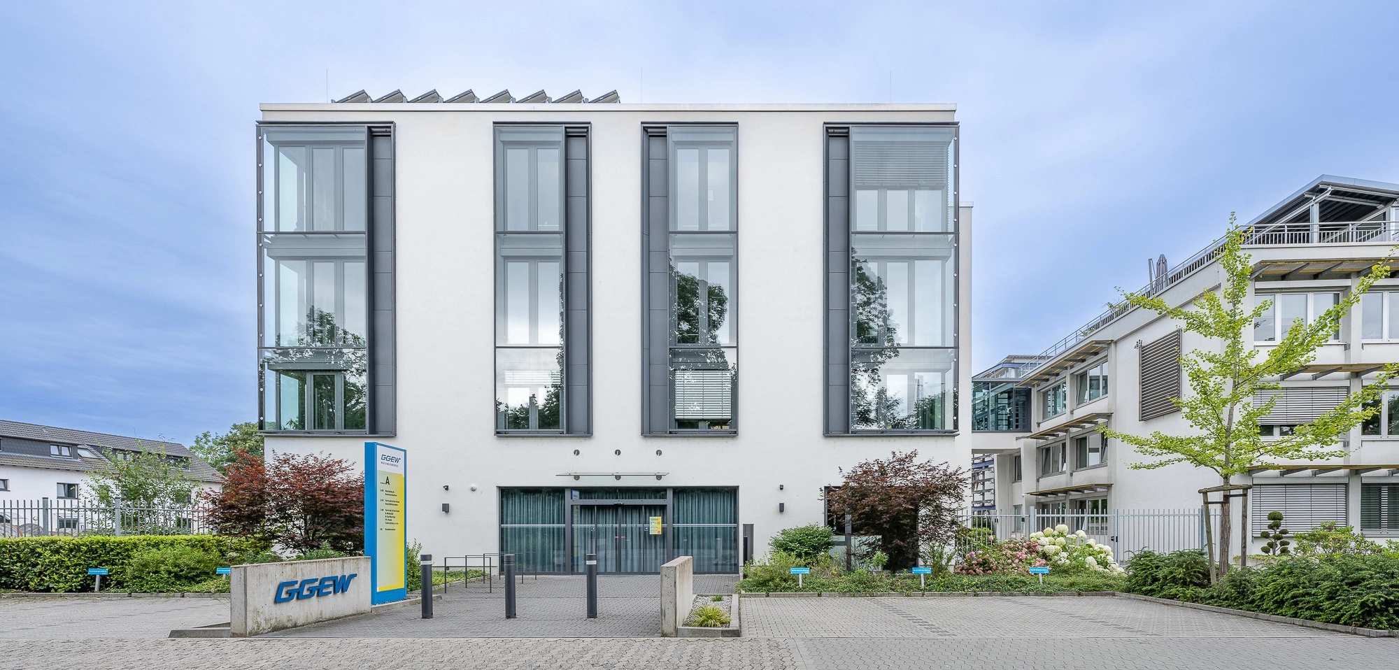 Modernes weißes Bürogebäude mit drei hohen Fensterachsen, Vorgarten und Eingangstür, umgeben von Büschen und Nebengebäuden.