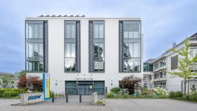 Modernes weißes Bürogebäude mit drei hohen Fensterachsen, Vorgarten und Eingangstür, umgeben von Büschen und Nebengebäuden.