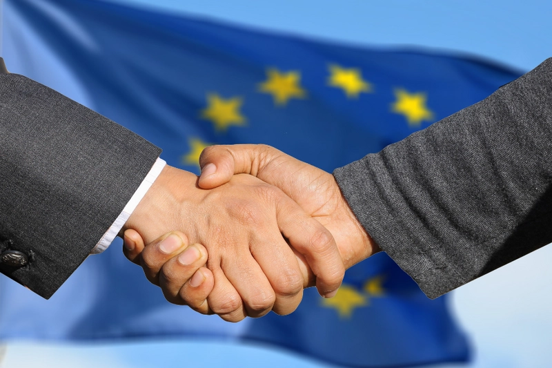 Zwei Hände schütteln sich vor einem unscharfen Hintergrund mit der Flagge der Europäischen Union. Die Geste symbolisiert Zusammenarbeit und Einigung.