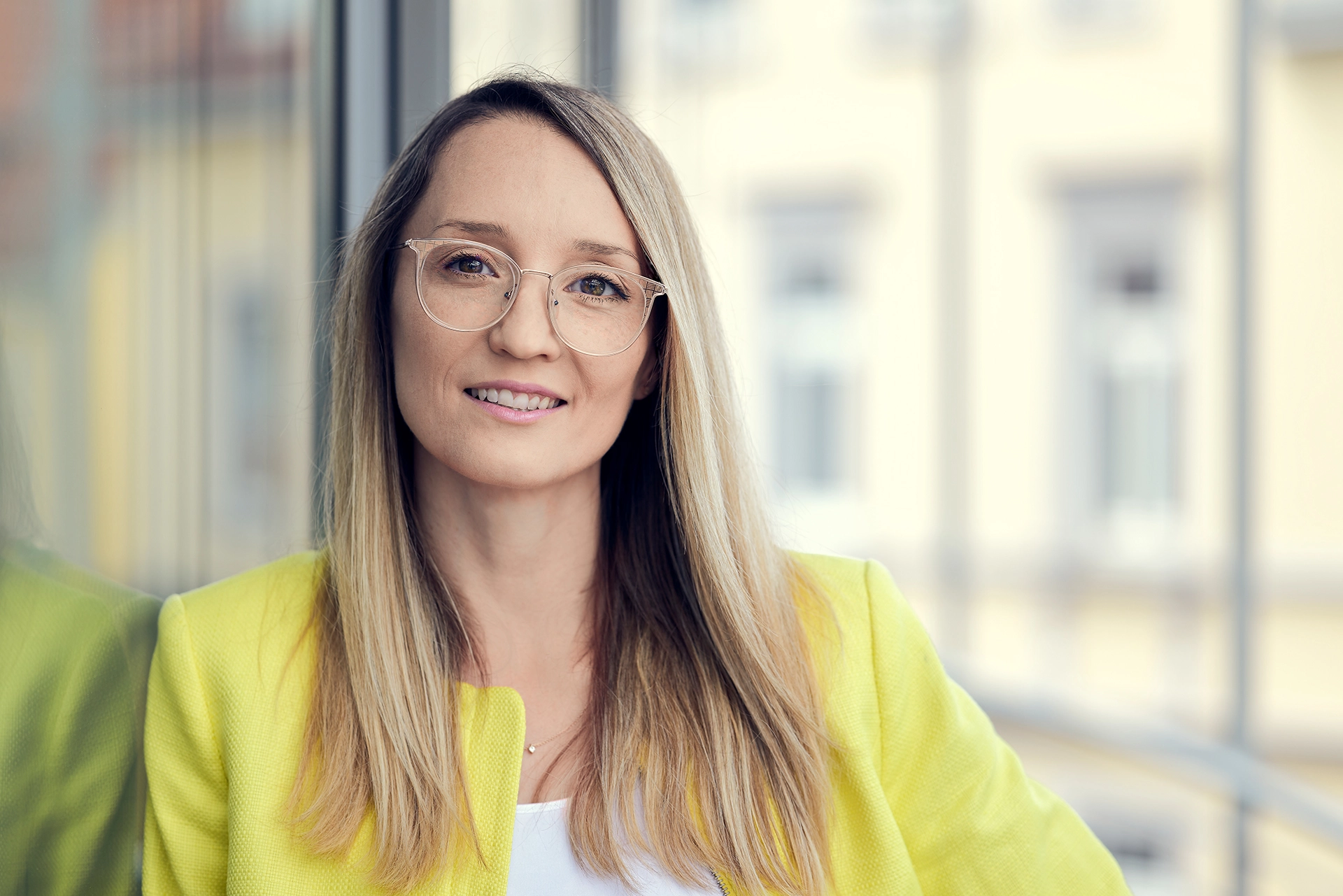 Porträt einer lächelnden Frau mit Brille und gelbem Blazer vor einem Fenster.