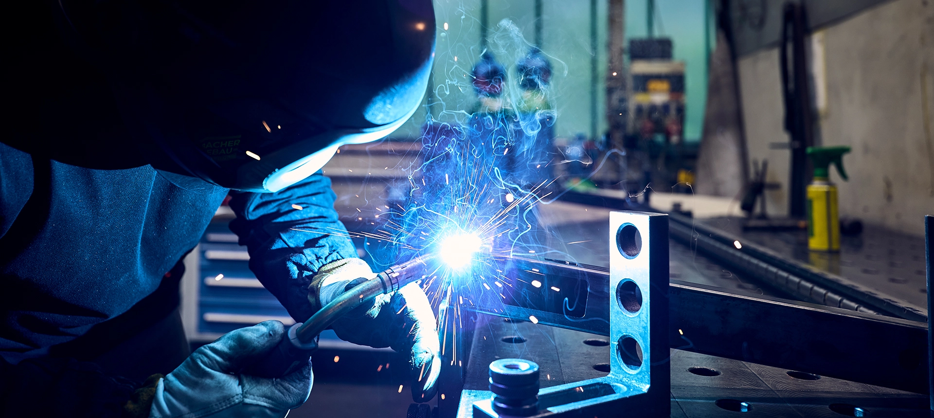 Person mit Schweißhelm und Handschuhen schweißt ein Metallteil; Funken sprühen.