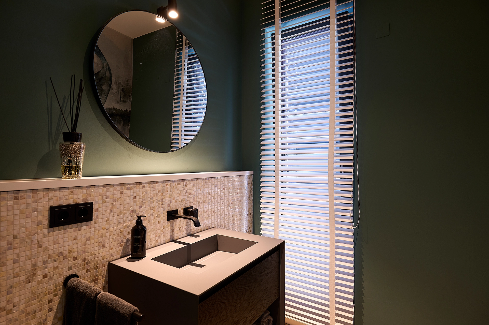 Modernes Badezimmer mit Waschtisch, Wandarmatur, rundem Spiegel und Fenster mit weißen Jalousien.