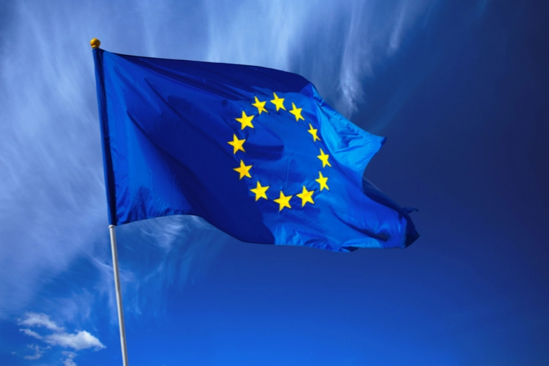 Eine blaue Flagge mit einem Kreis aus zwölf gelben Sternen in der Mitte, die die Europäische Union symbolisiert. Der Hintergrund zeigt einen klaren blauen Himmel mit einigen leichten Wolken.
