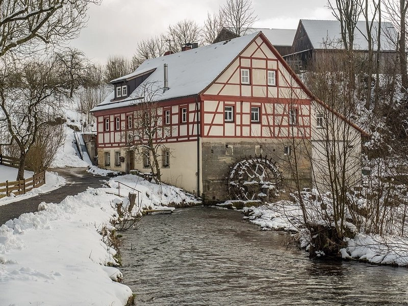 Ein historisches, zweigeschossiges Gebäude mit Fachwerkfassade steht an einem schneebedeckten Ufer. Neben dem Haus fließt ein ruhiger Wasserlauf, in dem ein Wasserrad sichtbar ist. Die Umgebung ist winterlich mit schneebedeckten Bäumen und einem grauen Himmel.