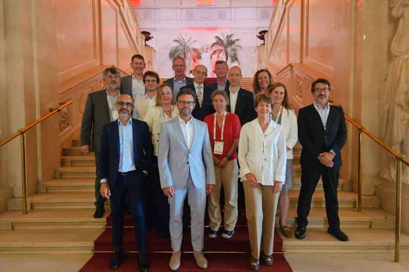Gruppensfoto von 15 Personen auf einer Treppe in einem eleganten Gebäude. Die Erwachsenen stehen in zwei Reihen, lächelnd und formell gekleidet. Im Hintergrund sind große Fenster und Palmen sichtbar.