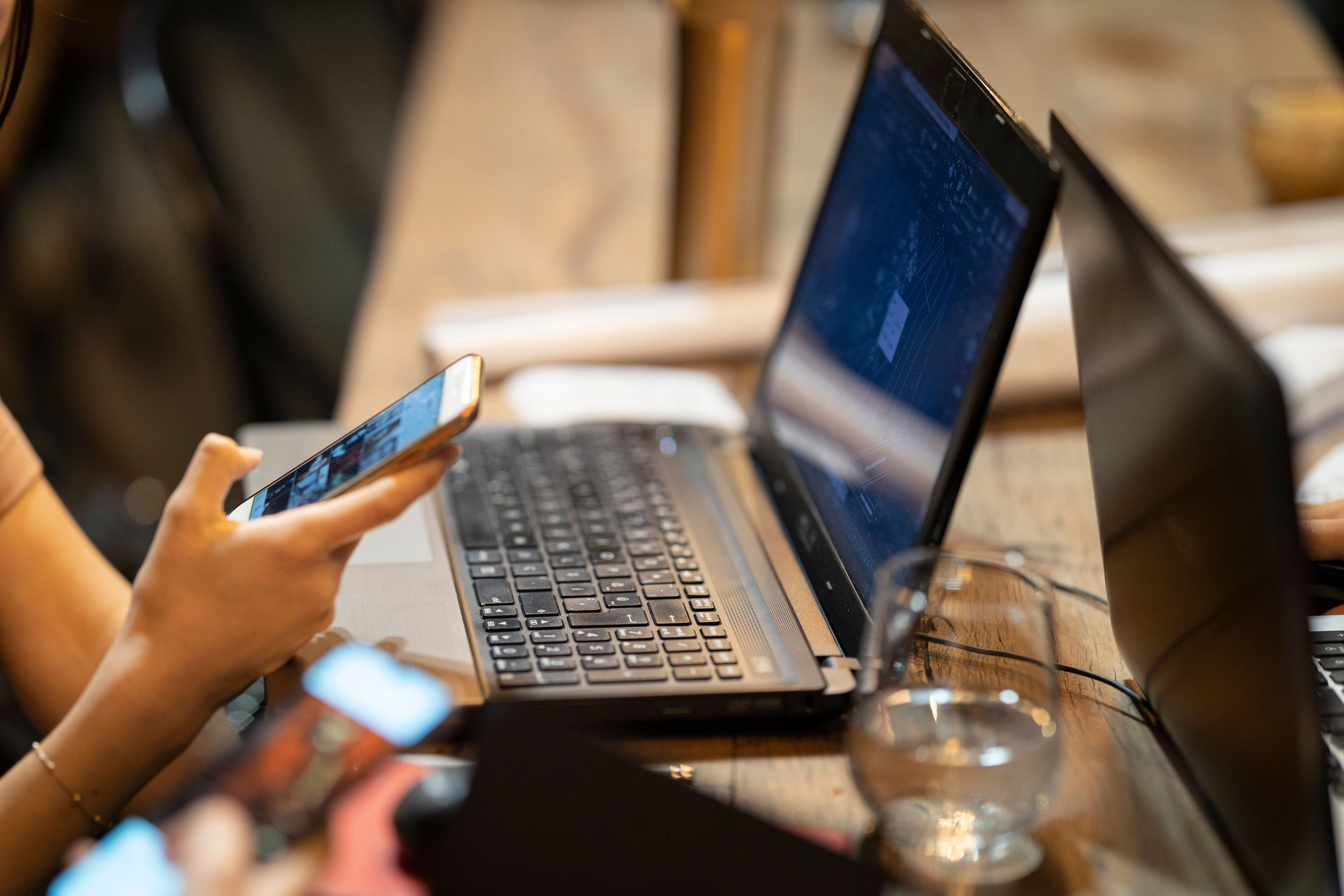 Nahaufnahme von Laptops und Händen, die Smartphones benutzen, auf einem Holztisch mit einem Wasserglas.