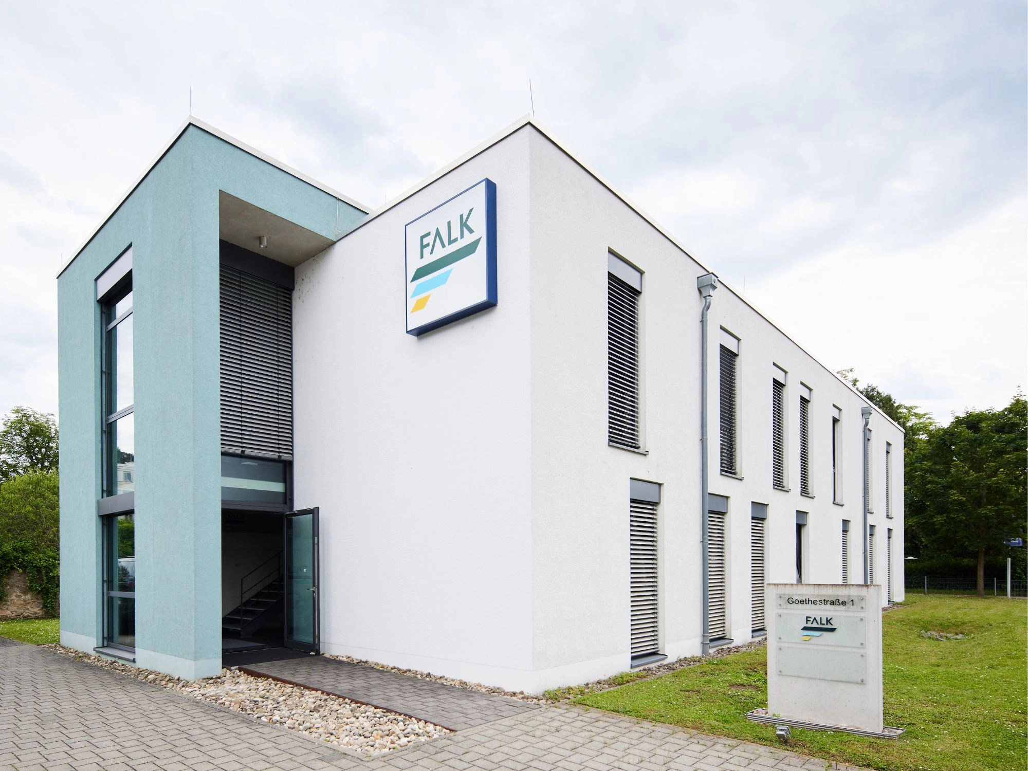 Zweistöckiges weißes Bürogebäude mit FALK-Logo an der Fassade, Eingangsbereich und Schildern auf dem Rasen.