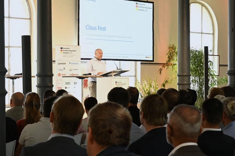 Ein Redner steht an einem Podium und spricht vor einem Publikum in einem hellen Raum mit großen Fenstern. Auf dem Bildschirm und im Hintergrund sind Präsentationsmaterialien zu sehen. Das Publikum, bestehend aus mehreren Personen, hört aufmerksam zu.