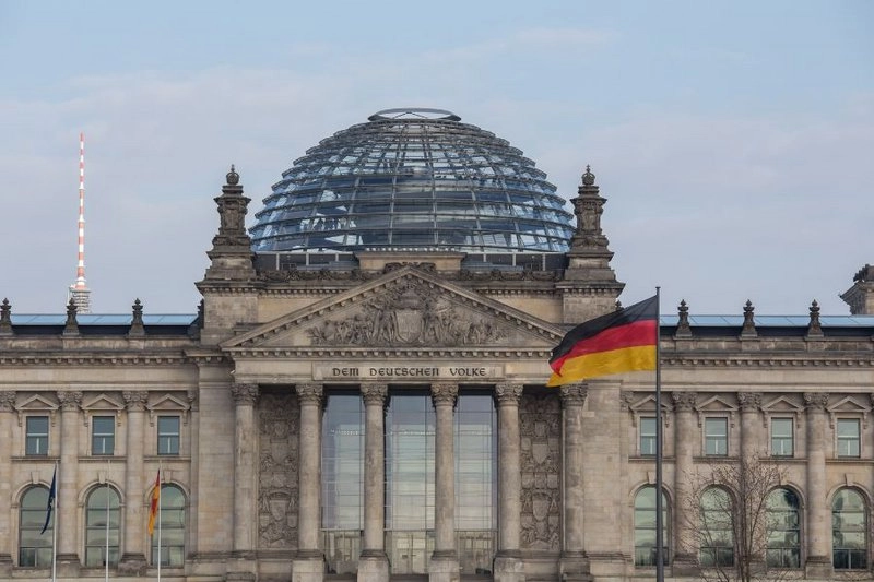 Das Bild zeigt die Fassade des Reichstagsgebäudes in Berlin mit einer markanten Glaskuppel. Vor dem Gebäude weht die deutsche Flagge. Es ist ein klarer Himmel im Hintergrund zu sehen.
