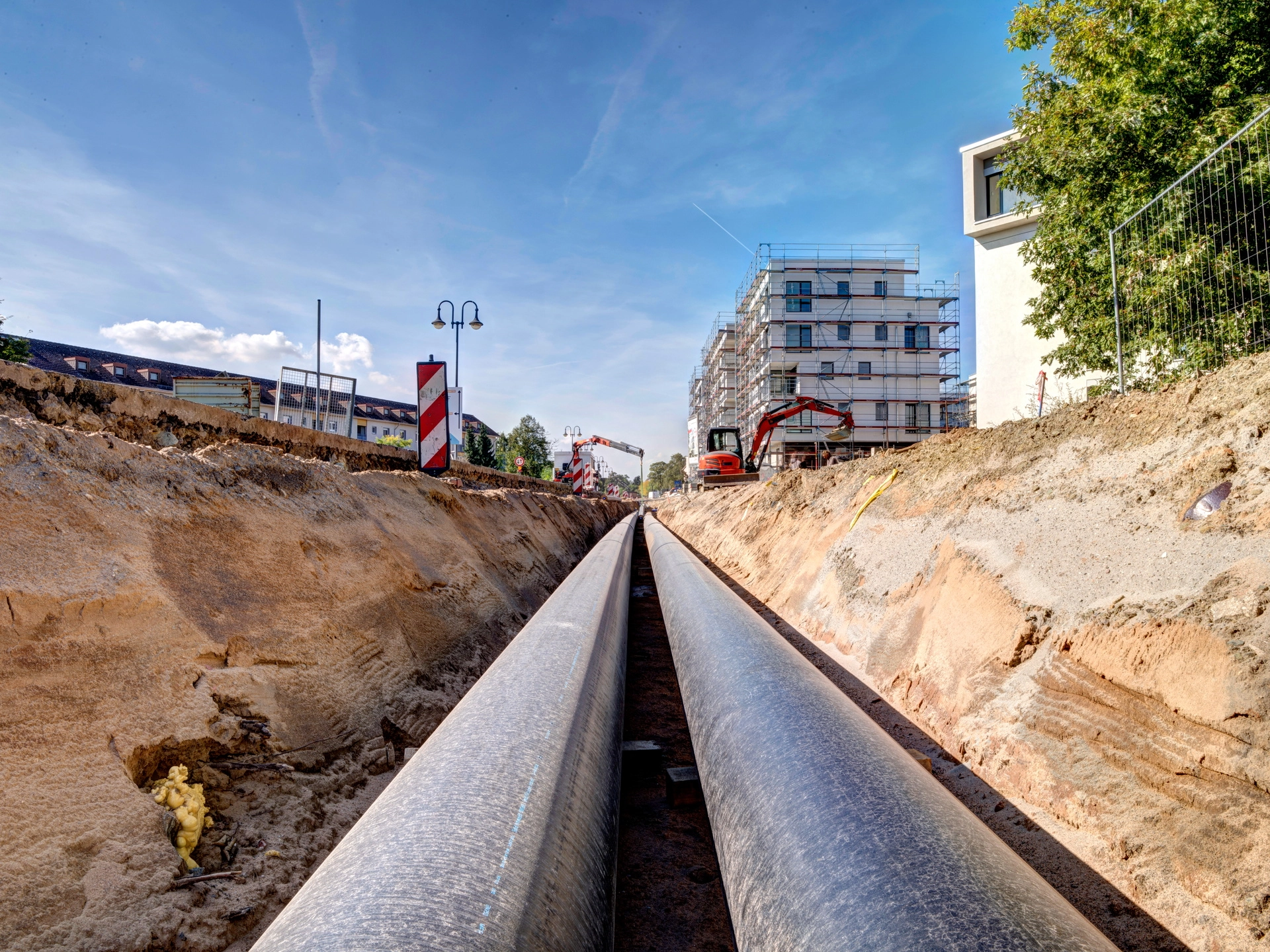 Ausgehobener Graben mit zwei parallel verlegten großen Rohrleitungen, Bagger und ein mehrstöckiges Gebäude im Hintergrund.