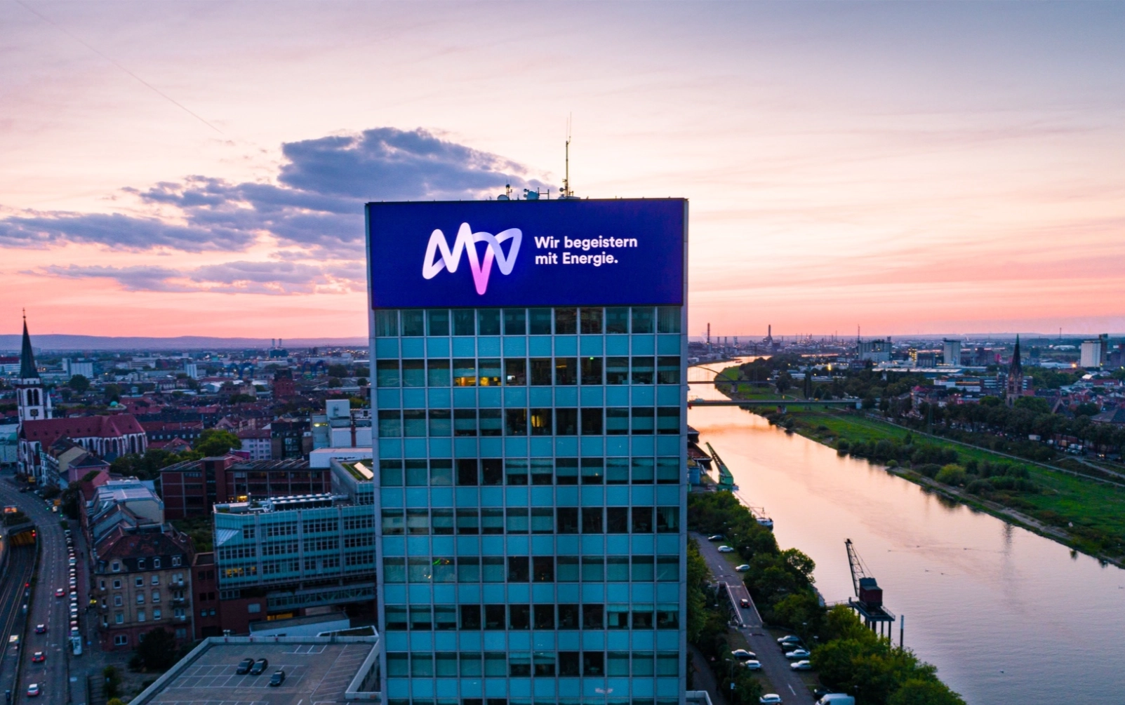 Hochhaus mit leuchtender Firmenwerbung an einem Flussufer, Stadt und Sonnenuntergang im Hintergrund.