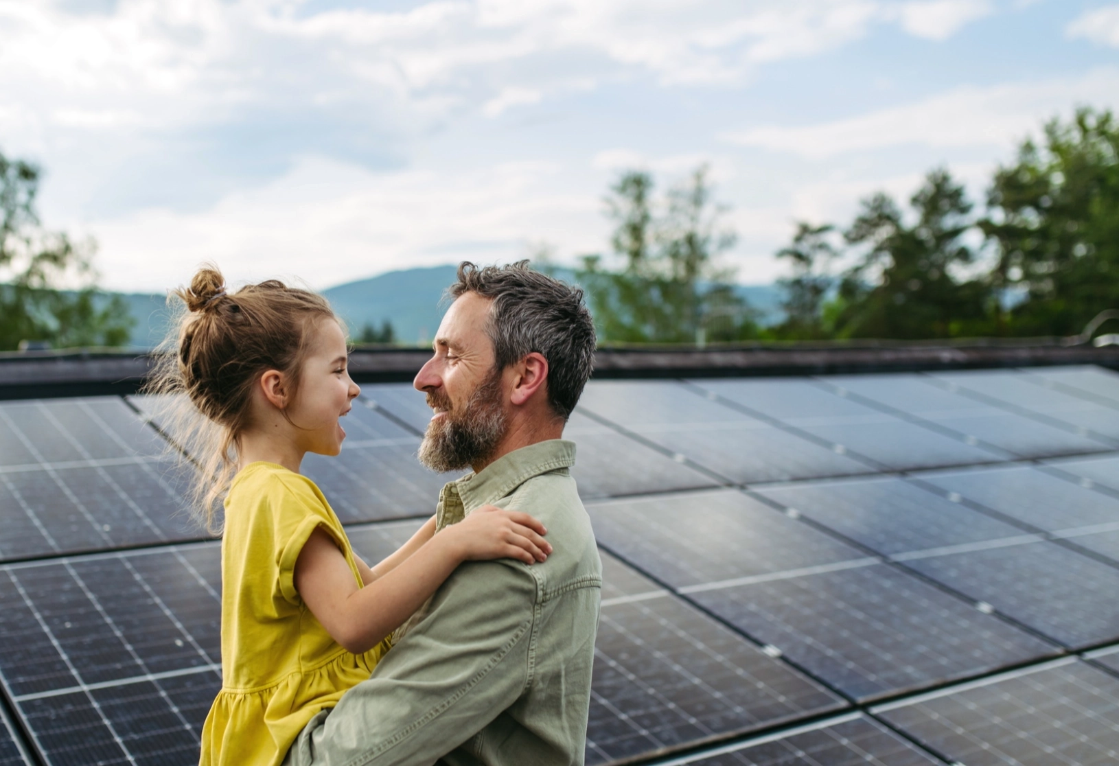 Ein Mann hält ein lächelndes kleines Mädchen vor einer Reihe Solarpaneele auf einem Dach.