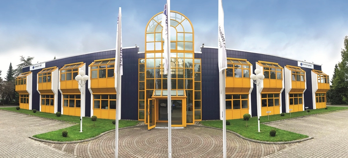 Zweistöckiges Bürogebäude mit gelben Fensterrahmen, großem Glaseingang und mehreren Fahnen im Vorfeld.