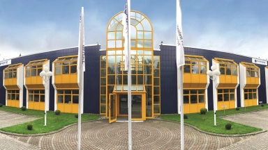 Zweistöckiges Bürogebäude mit gelben Fensterrahmen, großem Glaseingang und mehreren Fahnen im Vorfeld.