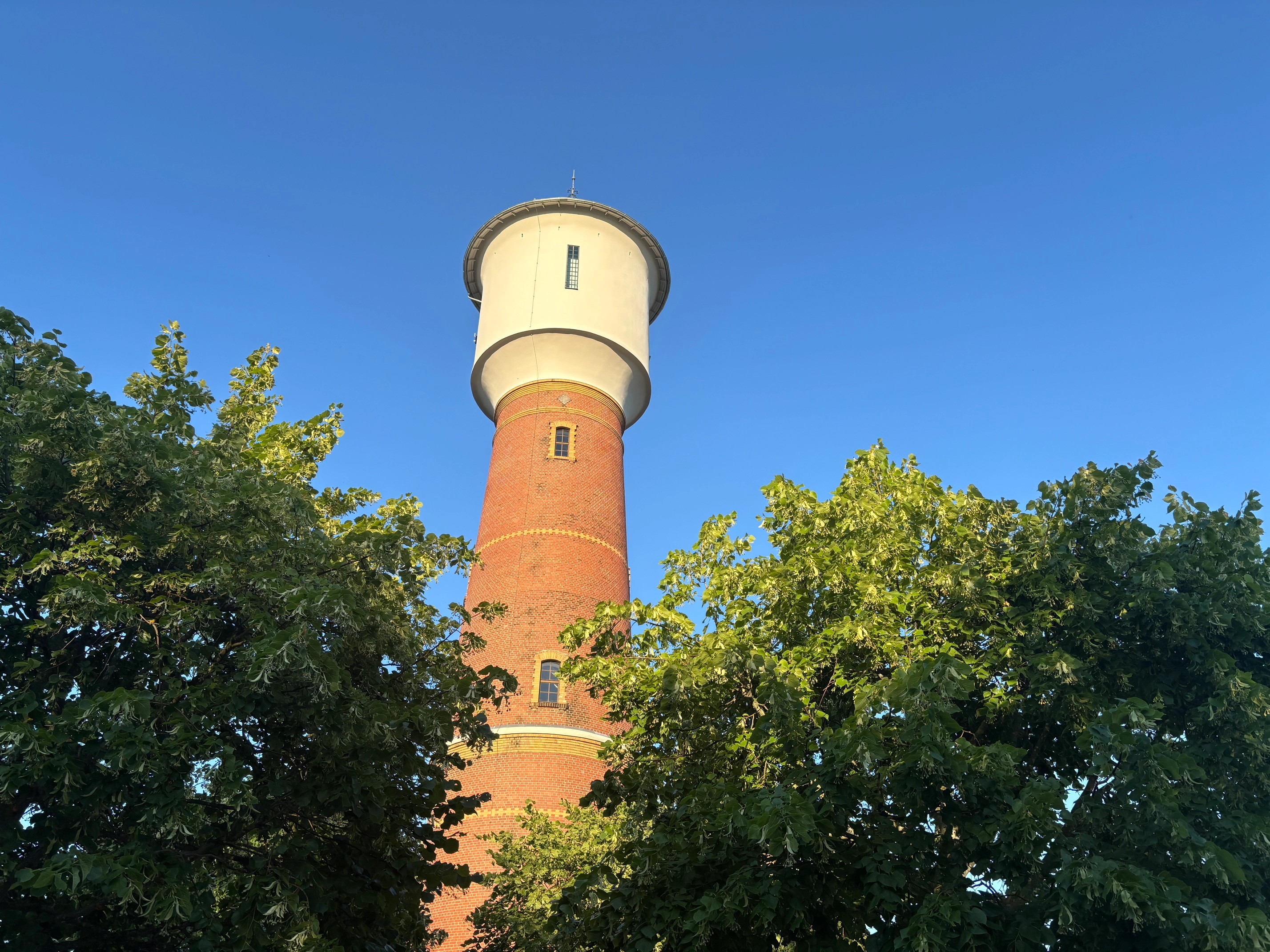 Roter Backstein-Wasserturm, vorne grüne Bäume, klarer blauer Himmel