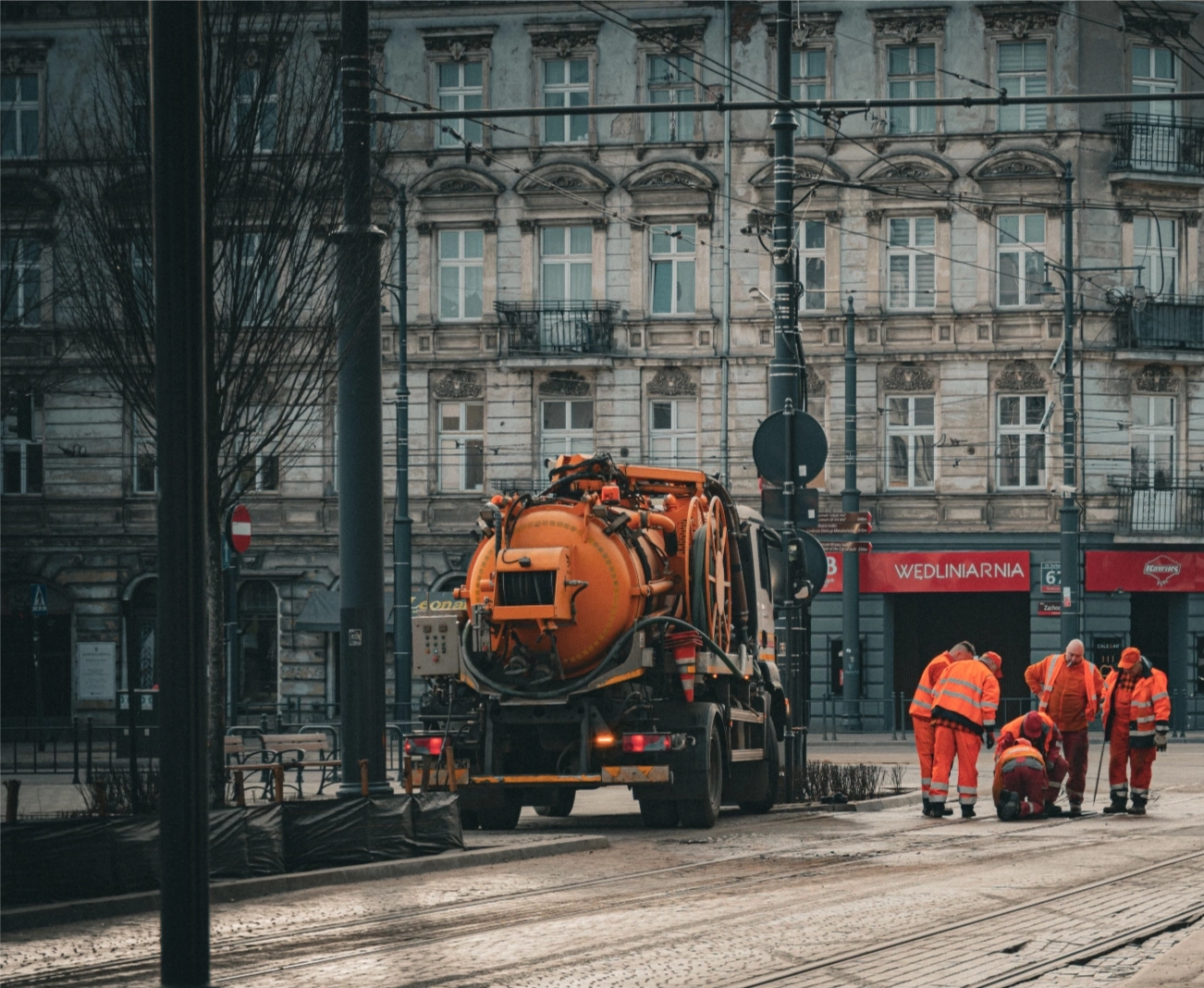 Orangefarbenes Wartungsfahrzeug und mehrere Arbeiter in orange Warnkleidung arbeiten auf einer Straße mit Straßenbahngleisen vor historischen Gebäuden.