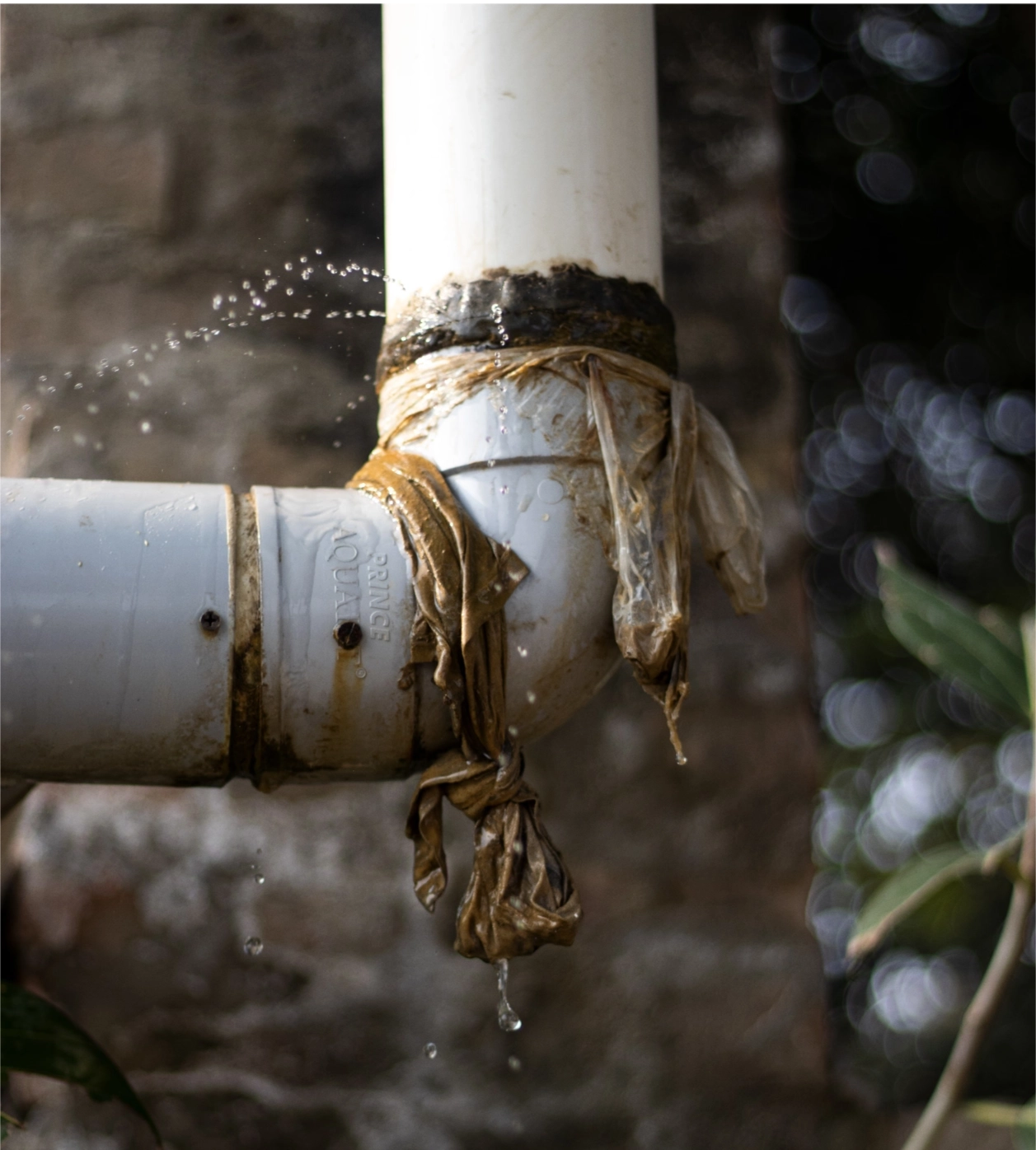 Nahaufnahme eines undichten weißen Abflussrohrs, an der Verbindungsstelle ist Plastik als provisorische Abdichtung befestigt und Wasser tropft heraus.