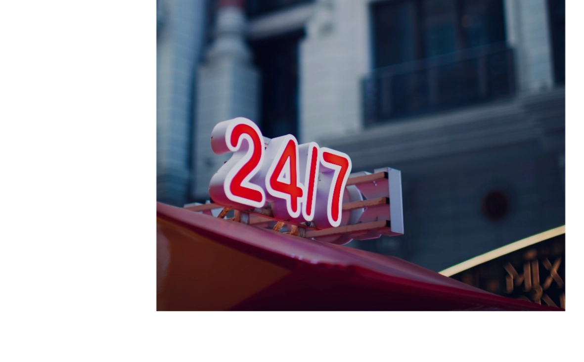 Rotes Leuchtschild mit weißer Umrandung und der Aufschrift „24/7“ an einer Straßenfassade.