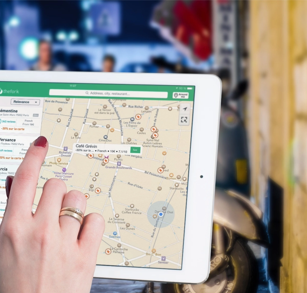 Hand mit zwei Ringen tippt auf ein Tablet, das eine Stadtkarte mit Restaurants und Suchfeld anzeigt.