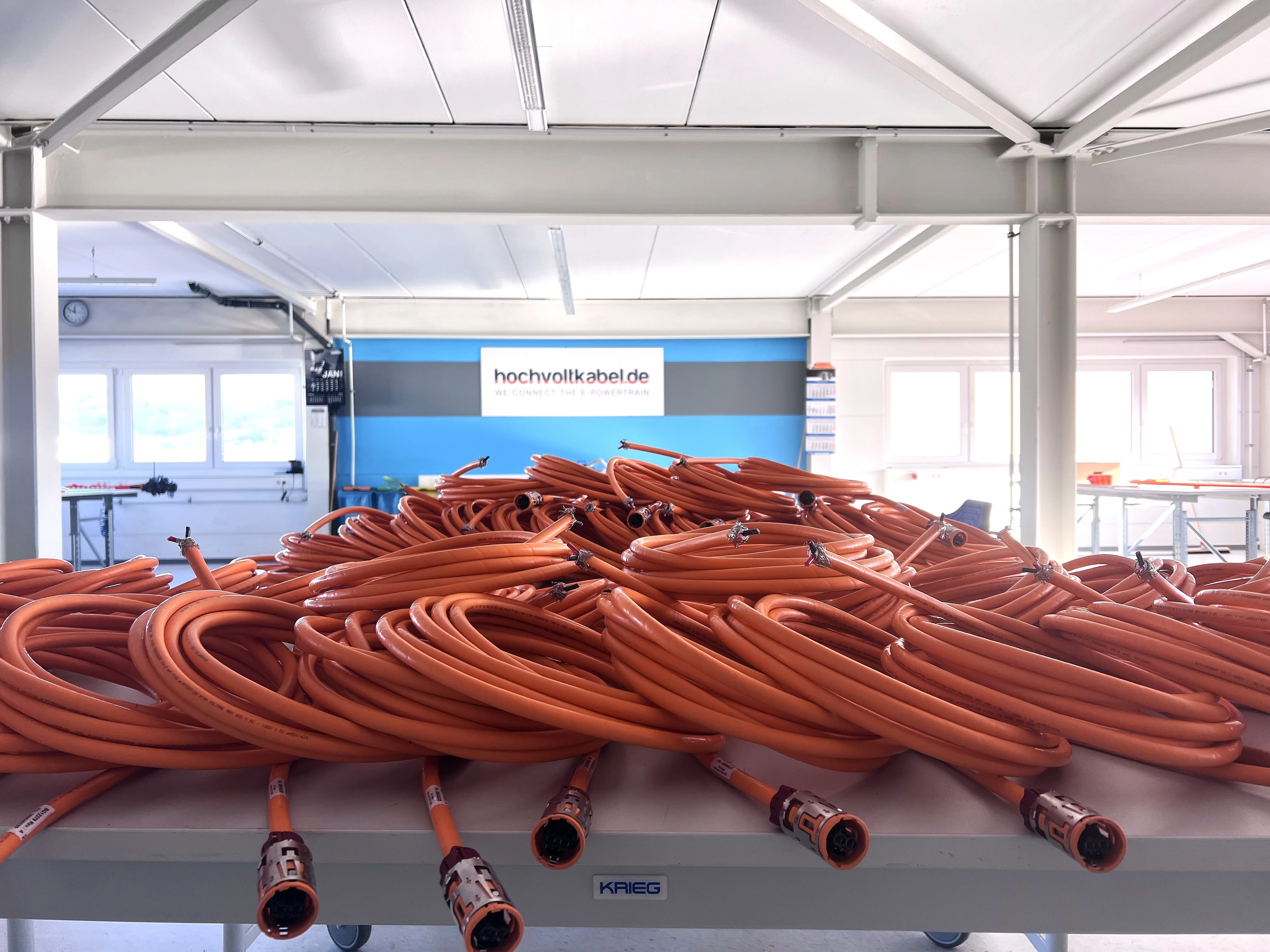 Viele orangefarbene Hochvoltkabel auf einem Tisch in einer hellen Werkhalle mit Werkstattumgebung im Hintergrund.