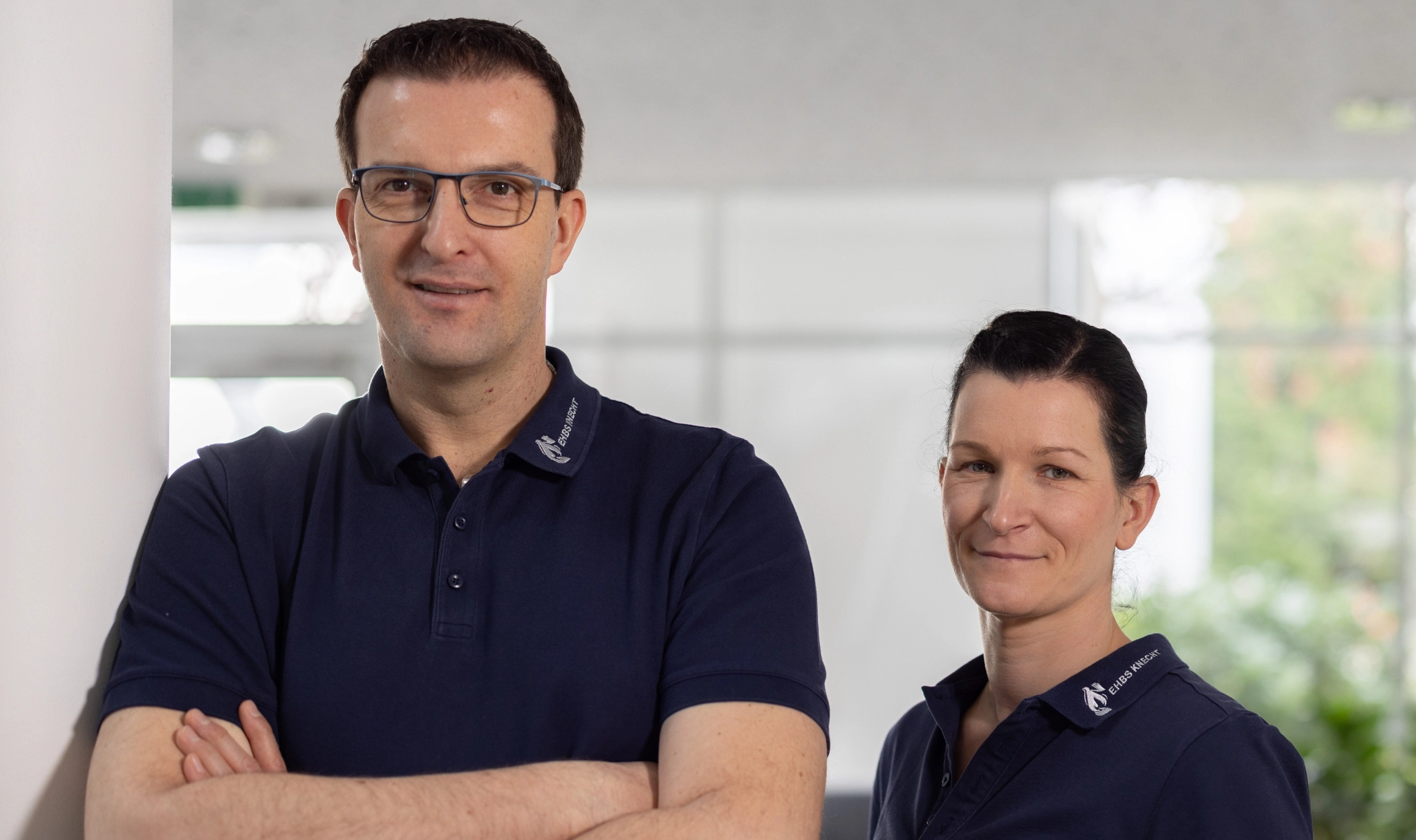 Zwei Personen in dunkelblauen Poloshirts stehen in einem hellen Innenraum; ein Mann mit verschränkten Armen vorne links und eine Frau leicht dahinter rechts.