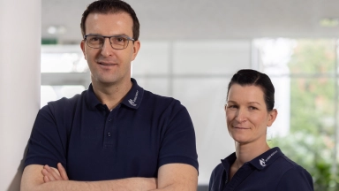 Zwei Personen in dunkelblauen Poloshirts stehen in einem hellen Innenraum; ein Mann mit verschränkten Armen vorne links und eine Frau leicht dahinter rechts.