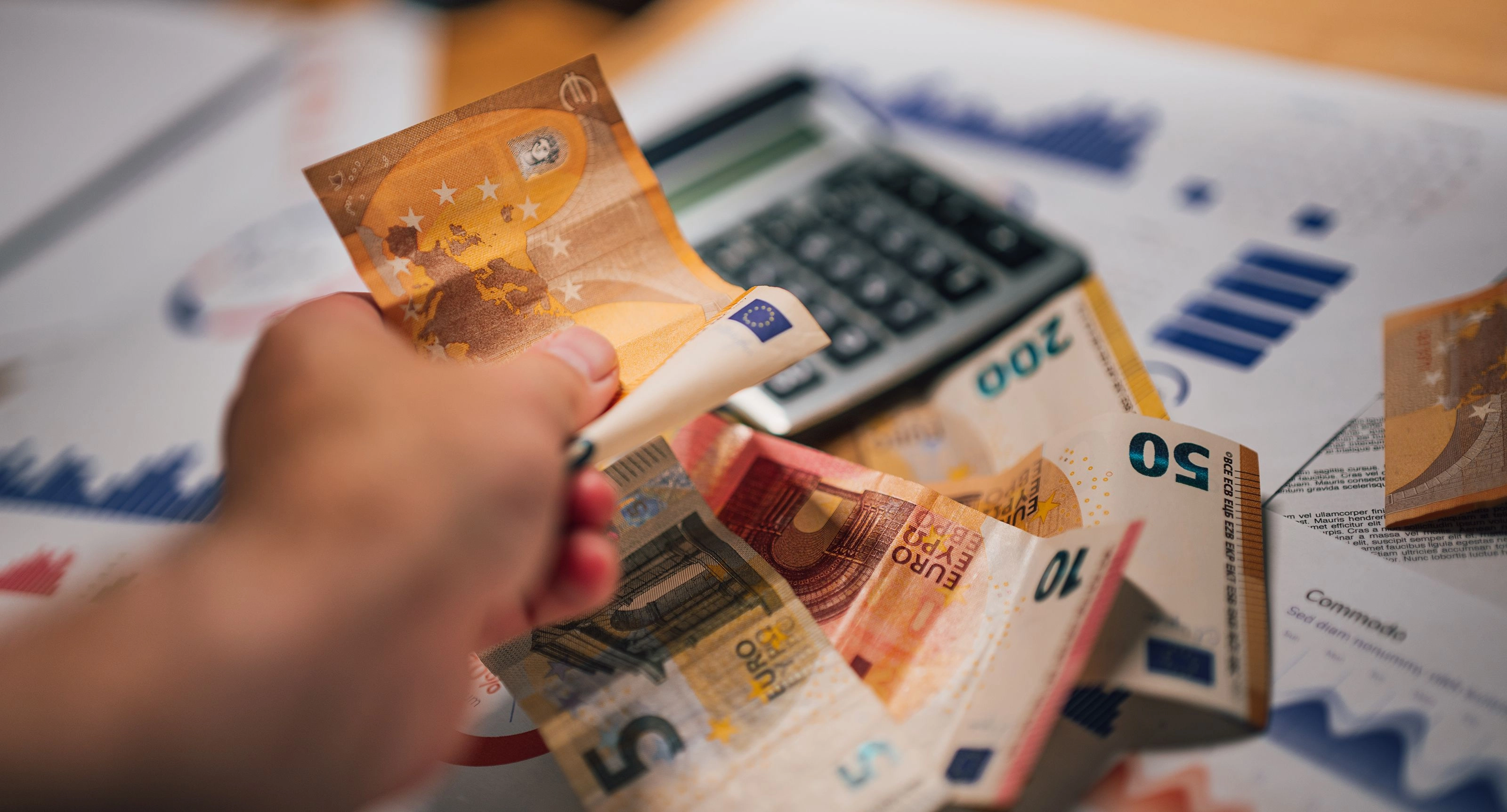 Hand, die mehrere Euro-Banknoten über einem Tisch mit Taschenrechner und gedruckten Finanzdiagrammen hält.