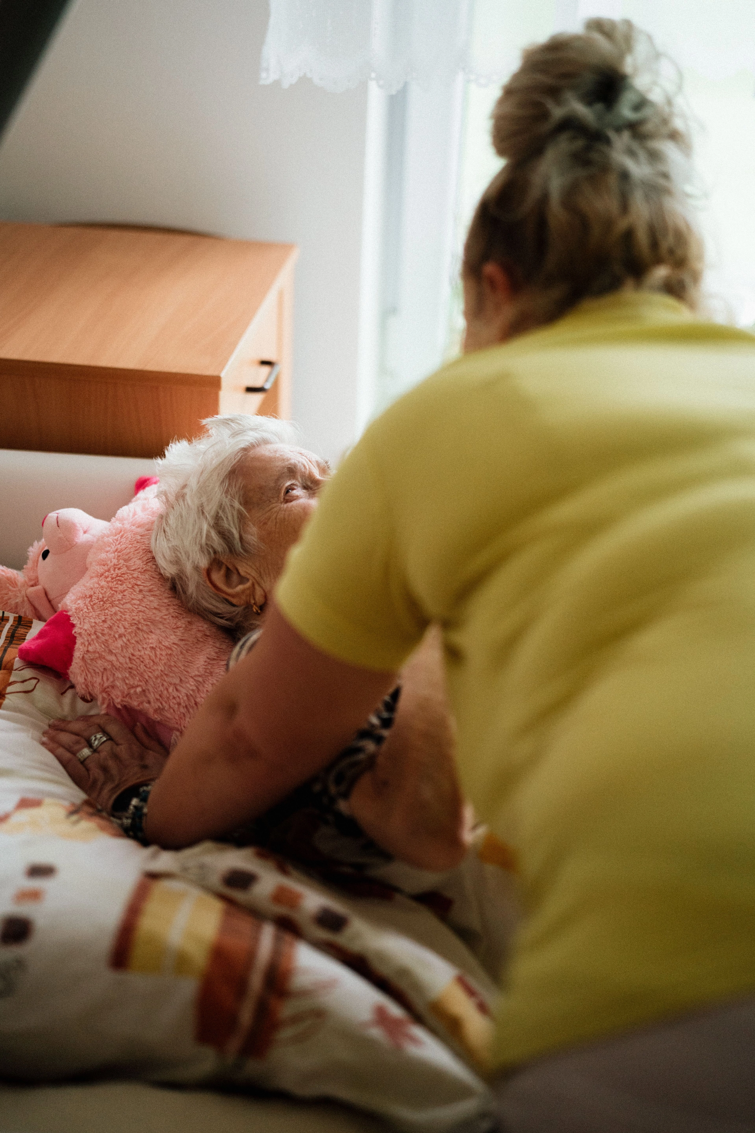 Ältere Frau liegt im Bett, wird von einer Pflegerin/Betreuerin umsorgt.