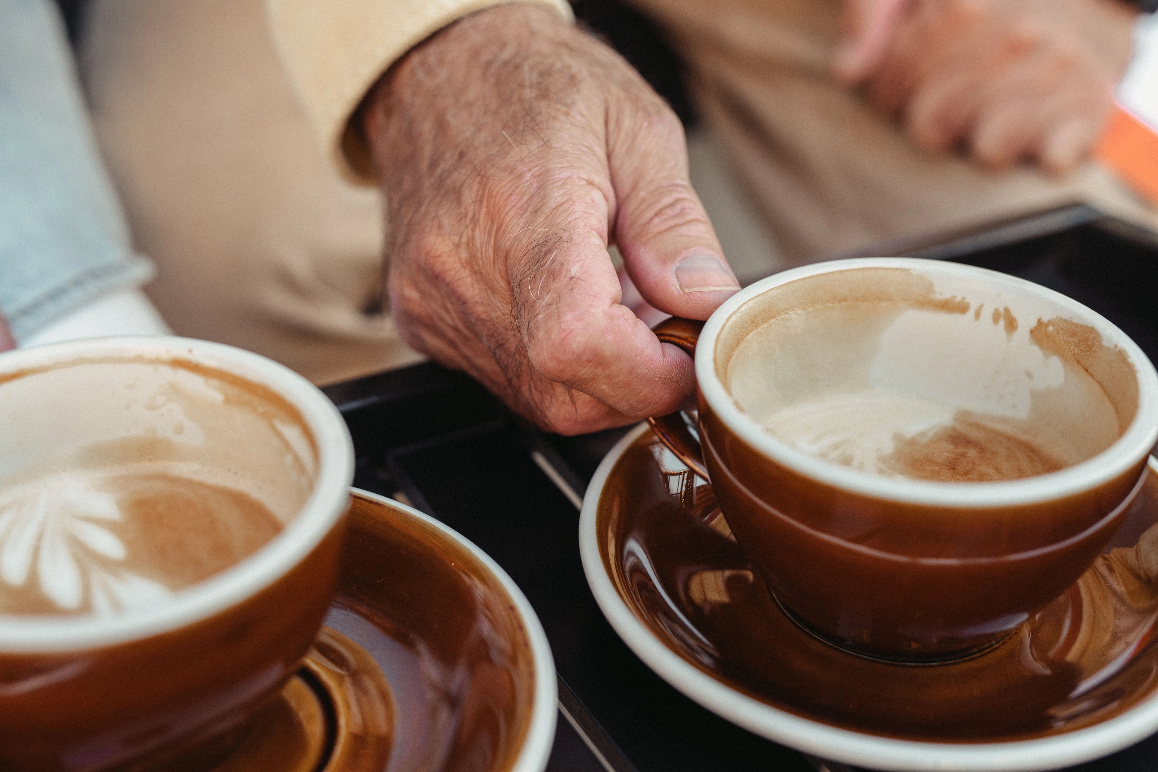 Nahaufnahme einer Hand, die eine braune Tasse mit Milchkaffee an einer Untertasse hält.
