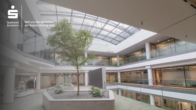 Innenraum eines modernen Atriums mit zentralem Pflanzbeet und Baum, umlaufenden Galerien und Glasdach; Sparkasse-Logo in der oberen Ecke.
